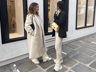 European influencer Amaka Hamelijnck and Bruna Rizk pose on a sidewalk in Paris wearing chic winter outfits with a long cream textured coat and a blazer with ribbed pants and sneakers.