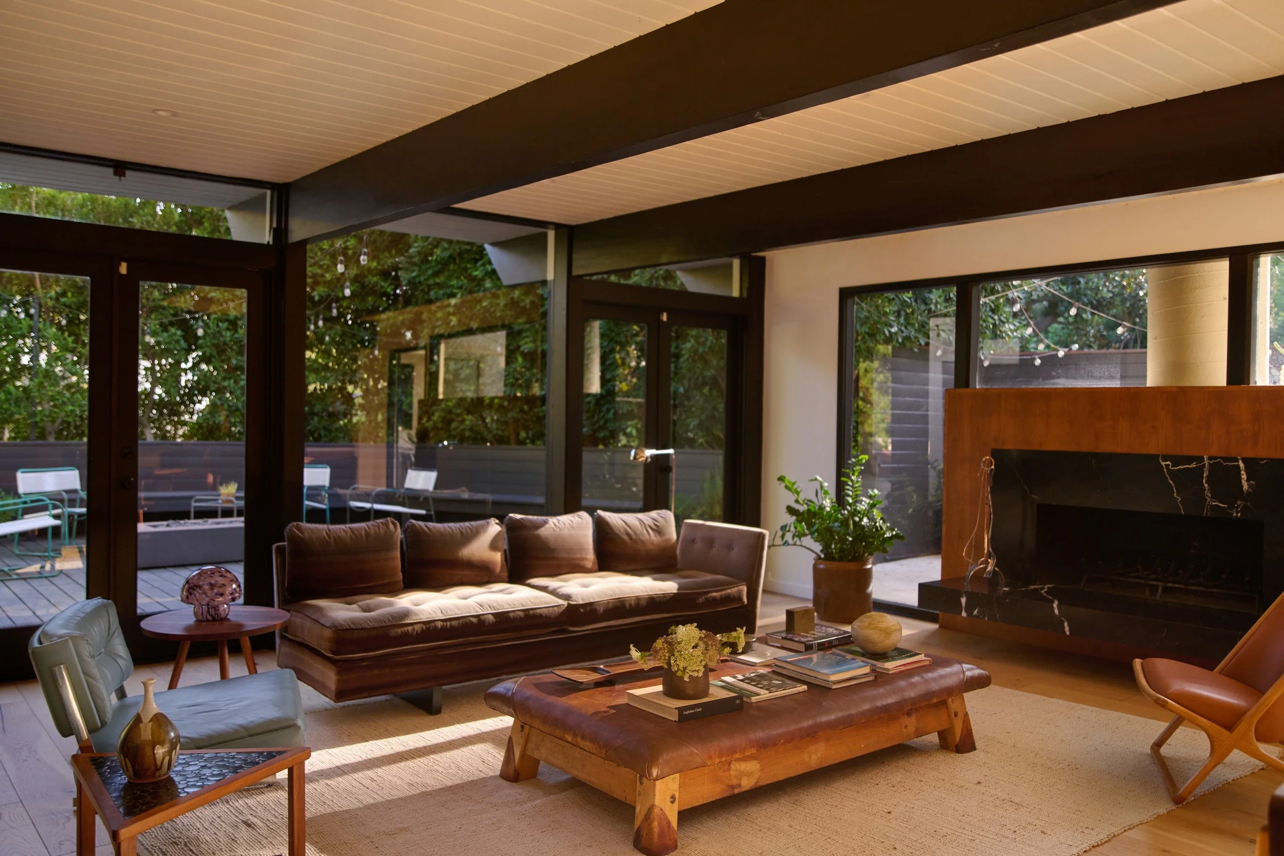 living room with one wall entirely glass windows and two doors. brown sofa, blue leather arm chair, wooden and brown leather coffee table on cream rug