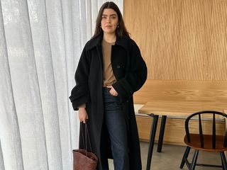 British fashion influencer Anna Newton poses in her modern wood kitchen in London wearing an oversize black coat, camel crewneck sweater, dark wash jeans, and a woven brown leather tote bag