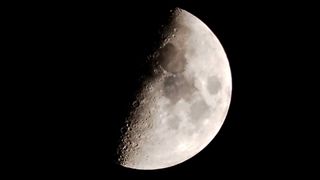 A picture of the moon against a black sky with its right side lit by direct sunlight and its left side bathed in shadow. Dark basaltic plains are visible marking the lunar surface, while numerous large craters can be seen sling the line separating night from day.