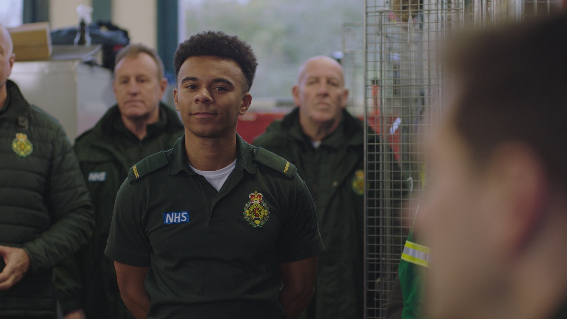 Teddy Gowan in his green paramedic uniform with his hands behind his back and smiling at an out of shot Iain Dean.