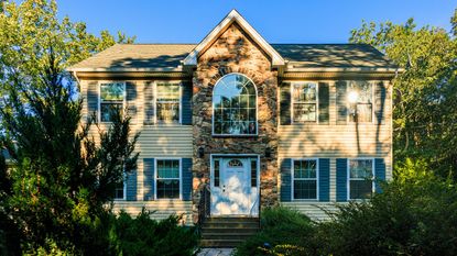 Two-story suburban house with distinctive stone facade and arched window. Beige siding, blue shutters, and green roof harmonize with natural surroundings. Lush greenery frames property, mature trees and shrubs add privacy. Stone steps lead to white front door, creating inviting entrance. 