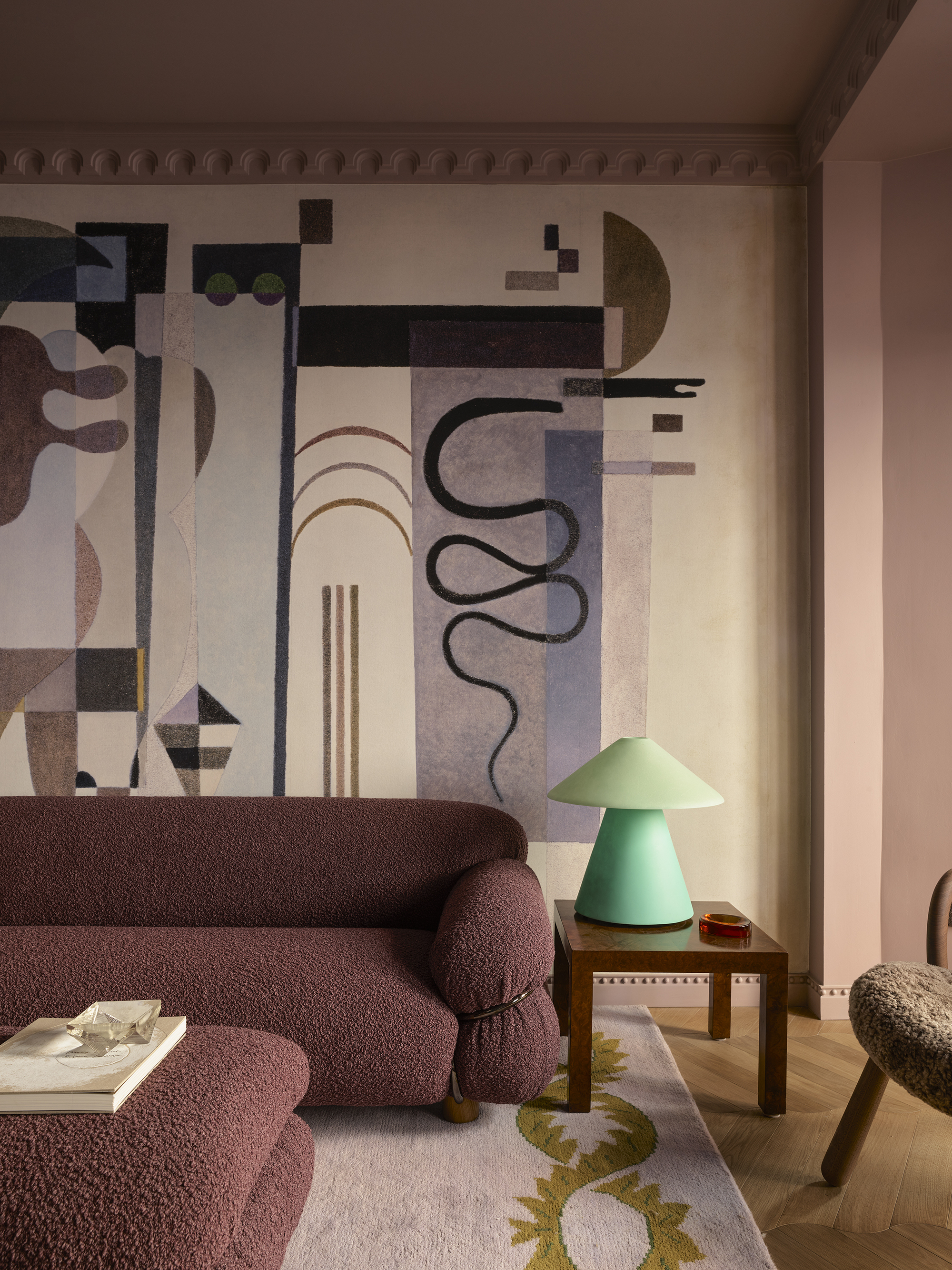 modern living room with pink walls, large textured wall mural, purple boucle sofa with matching ottoman coffee table, pale pink rug with mustard trim, herringbone floorboards, timber side table with neon green table lamp