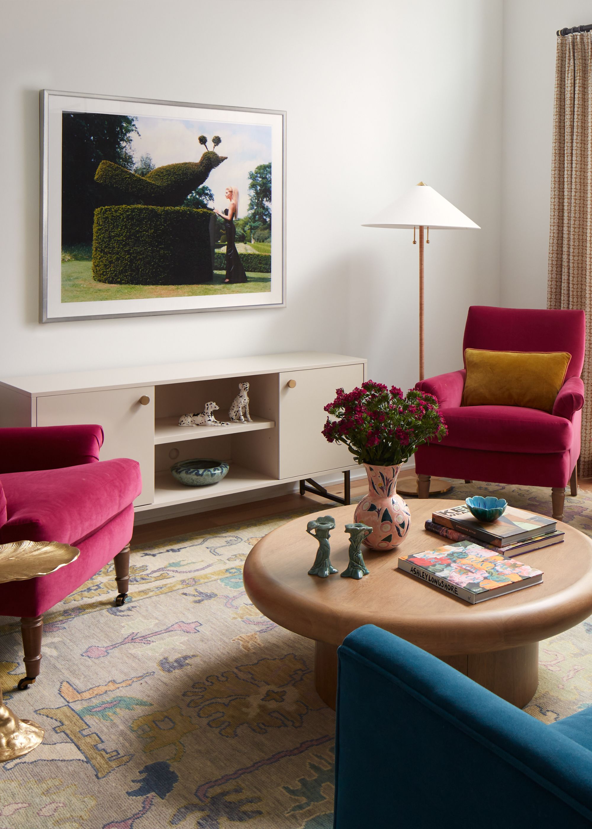 A modern living room with white walls, a beige patterned rug, a round wooden coffee table, and a sideboard with a piece of artwork above. Two hot pink armchairs and a dark blue sofa in the corner.