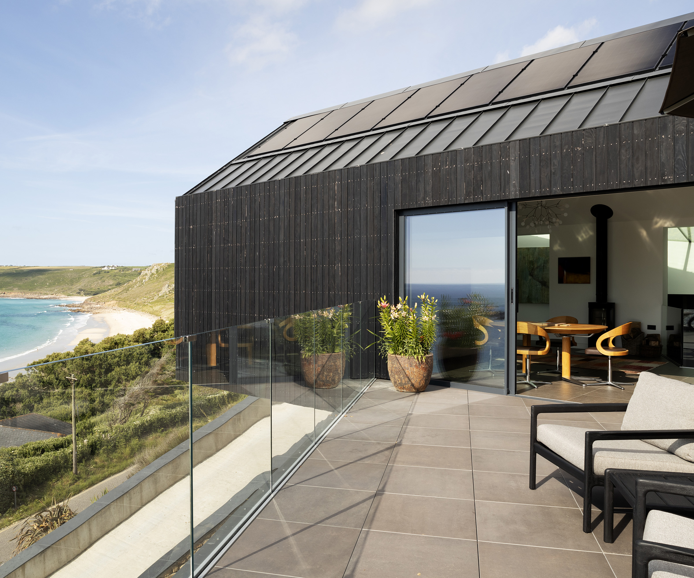 A sunny terrace next to a black timber clad house with sea views and glass ballustrade