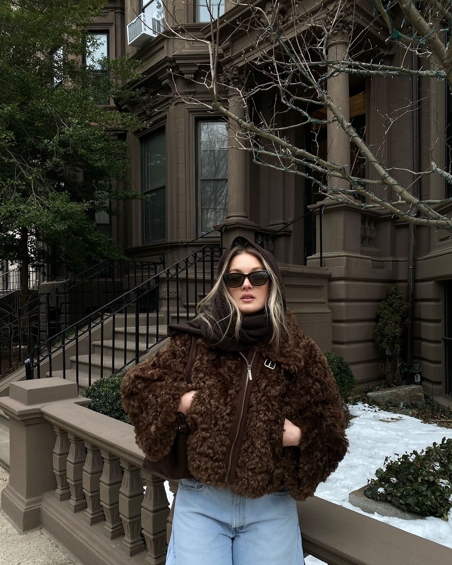 Uma mulher enrolada em um casaco marrom fofo do lado de fora de uma casa marrom nevada cercada por árvores