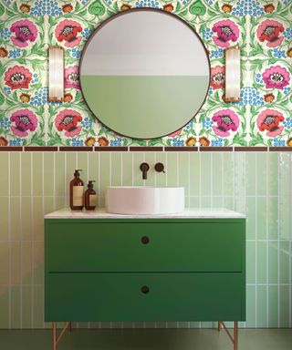 A green small bathroom with pink floral wallpaper, light green subway tile splashback, a dark green vanity unit, and a round mirror on the wall