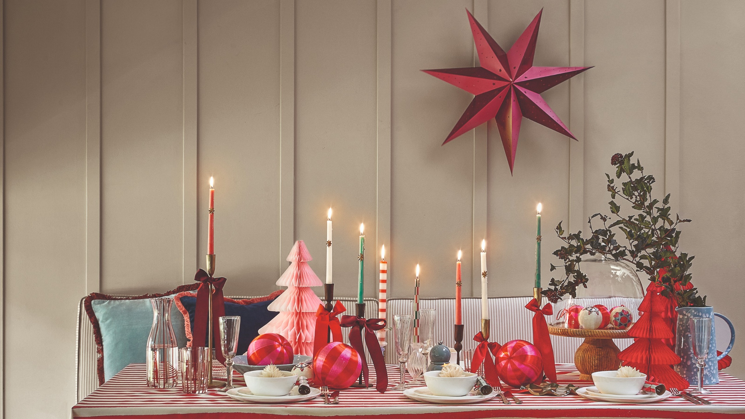 A Christmas dining table with lit dinner candles decorated with ribbons and a striped paper Christmas star decoration hung on the wall 