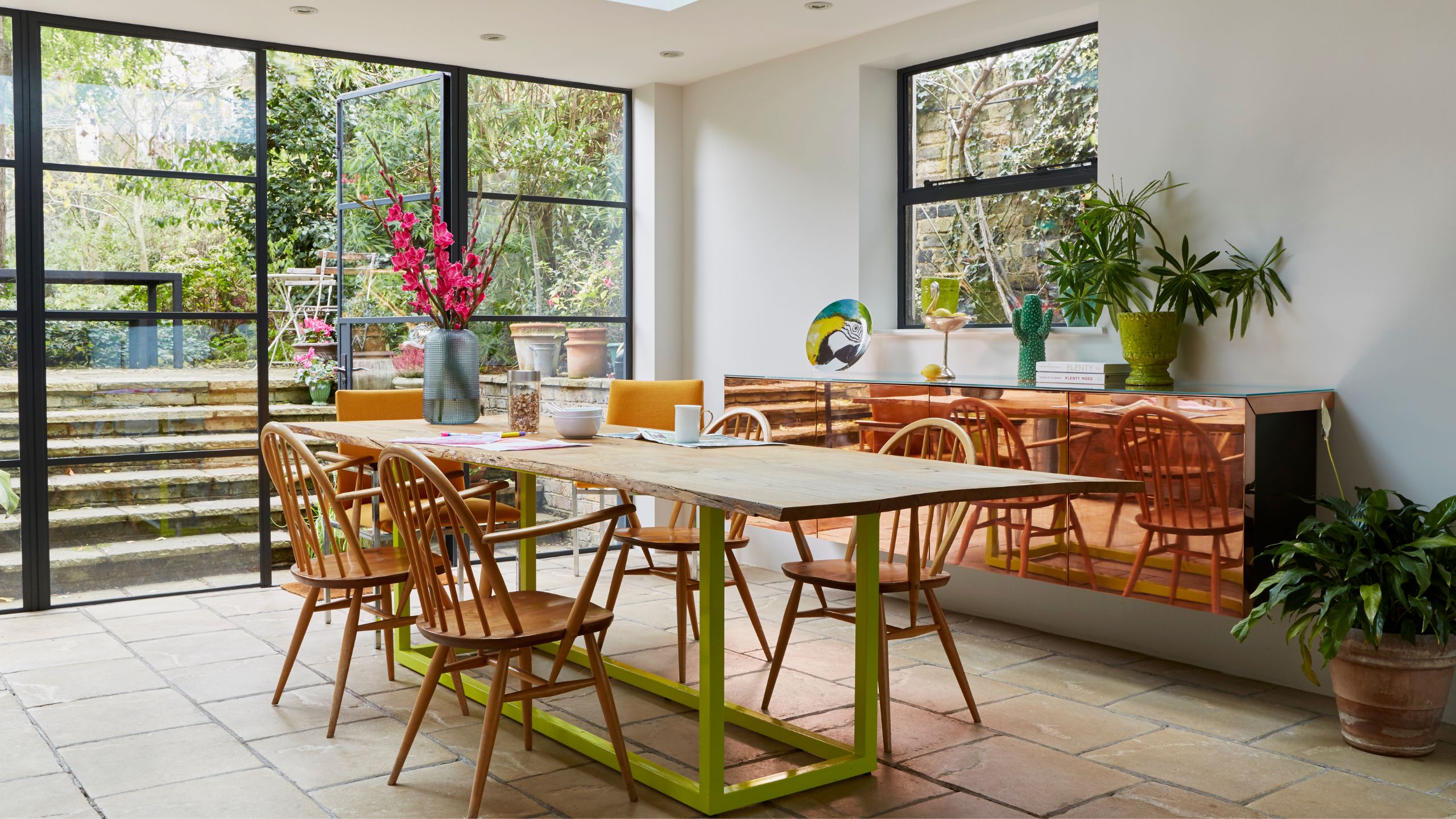 A modern kitchen extension with a rustic wood tabletop and lime green metal frame legs with round-backed wood dining chairs, and a reflective copper wall-hung console next to large glass doors