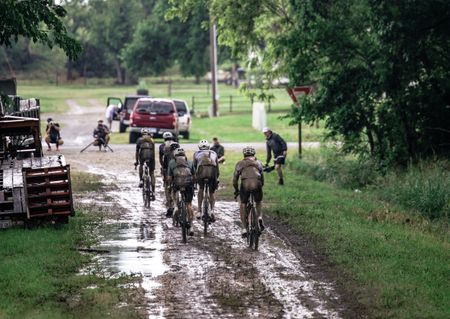 Elite riders march through the mud as they approach an paved intersection on the Unbound Gravel 200 course, with media present in the area
