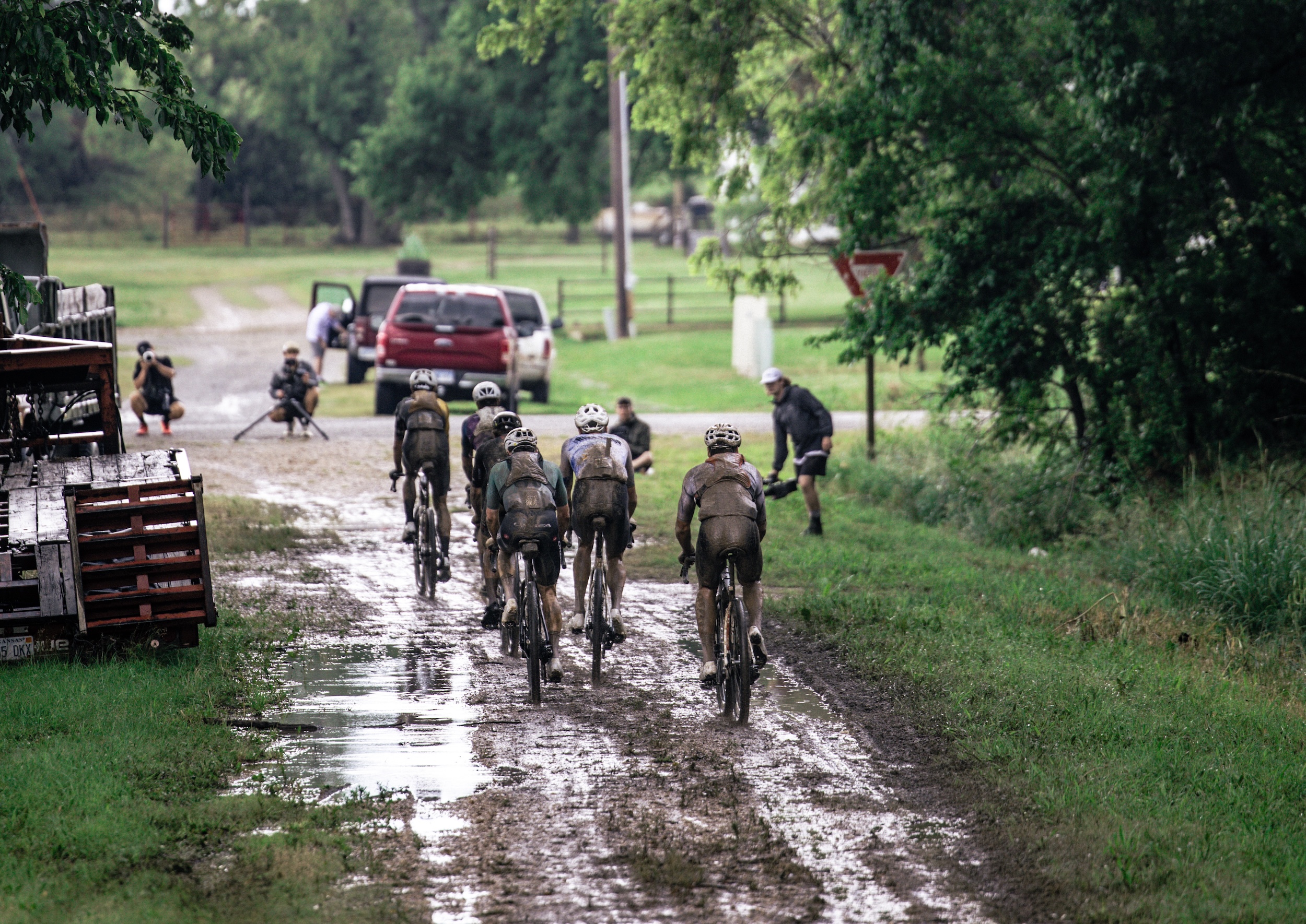 Elite riders march through the mud as they approach an paved intersection on the Unbound Gravel 200 course, with media present in the area
