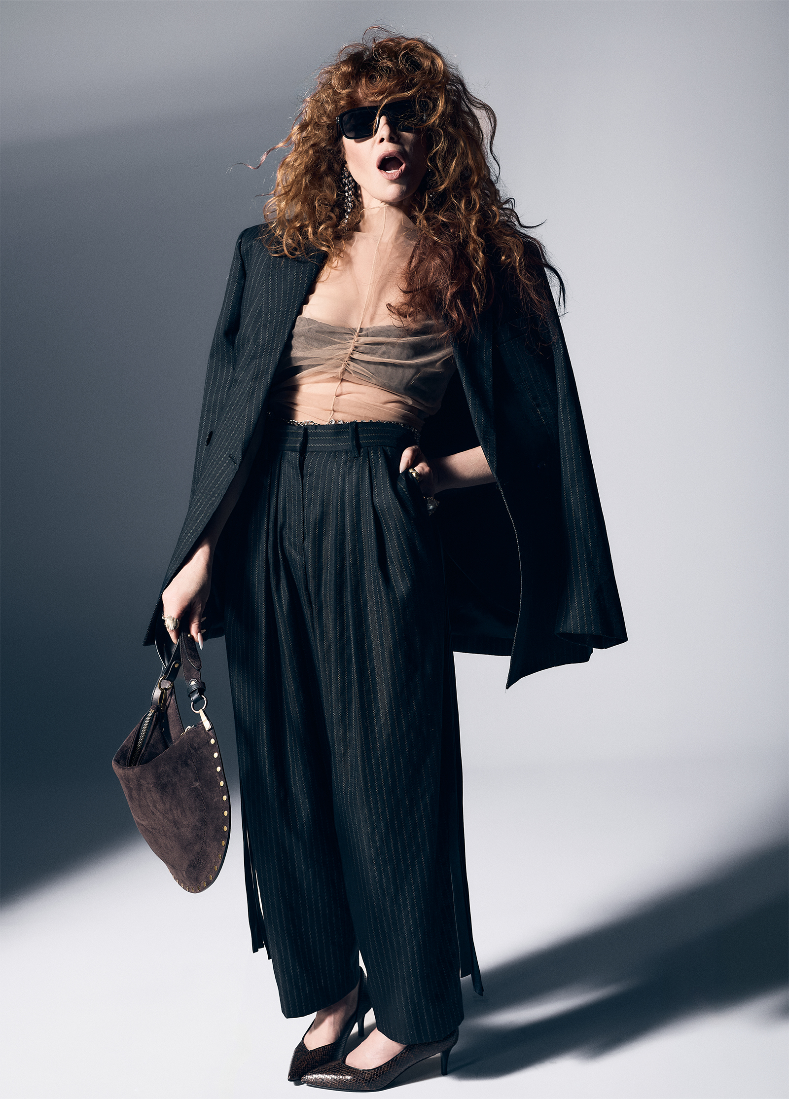 Natasha Lyonne photographed by Andrew Arthur for Who What Wear. She is is shot with her mouth open wearing an oversize pinstripe blazer and trousers with a nude sheer top and a brown suede bag.