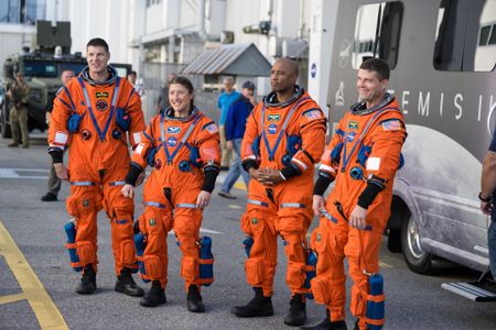 Four people in orange spacesuits stand on a road