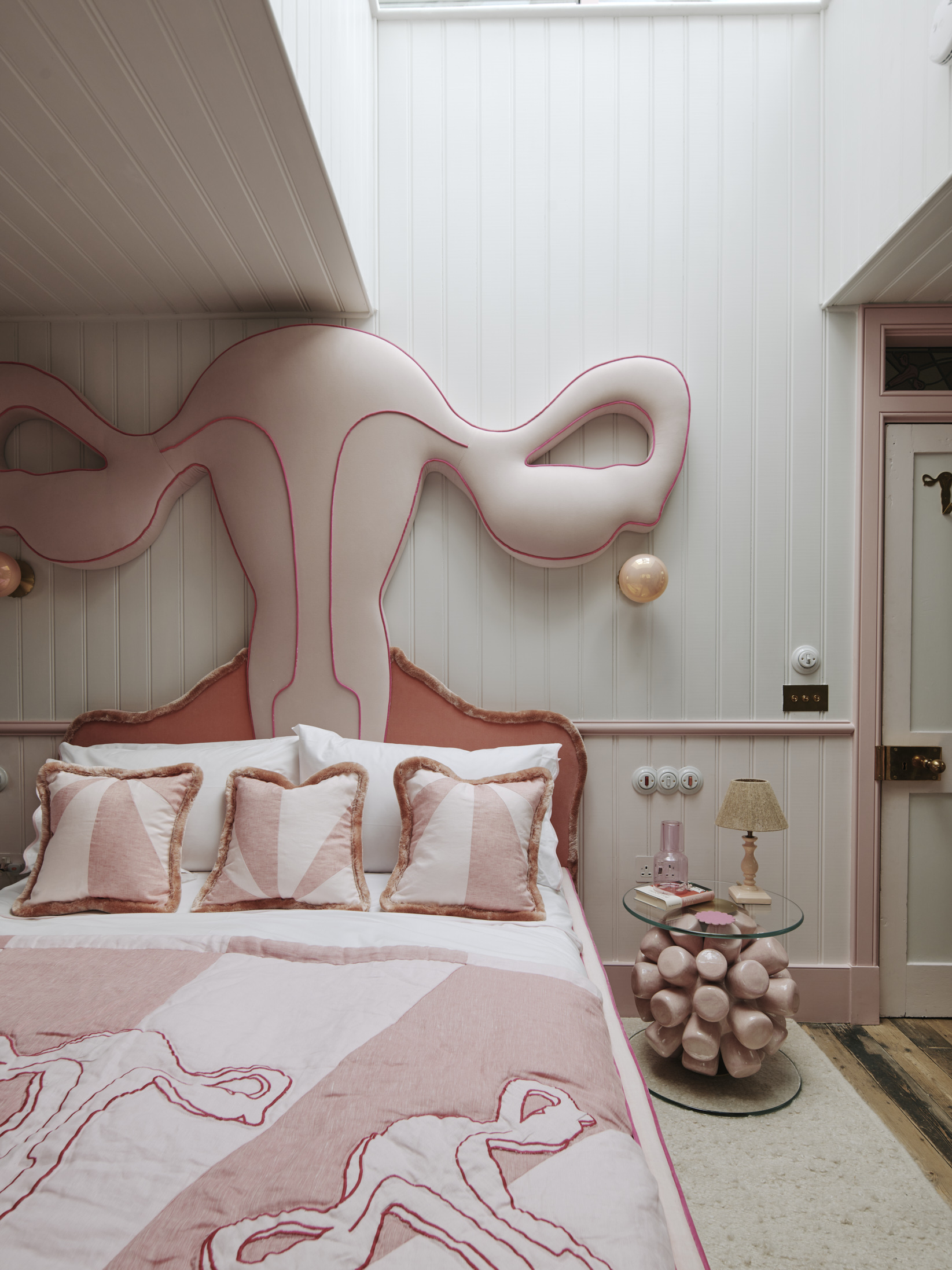 A plush pink bed with a headboard shaped like a uterus. Beside the bed is a night-table shaped like a ball of marshmallows.