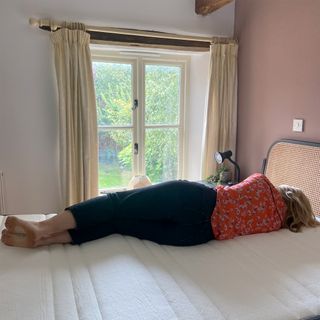 A woman with blonde hair wearing a red floral top and black jeans lying on her side on a mattress facing a window with a garden beyond. Her back is to the camera