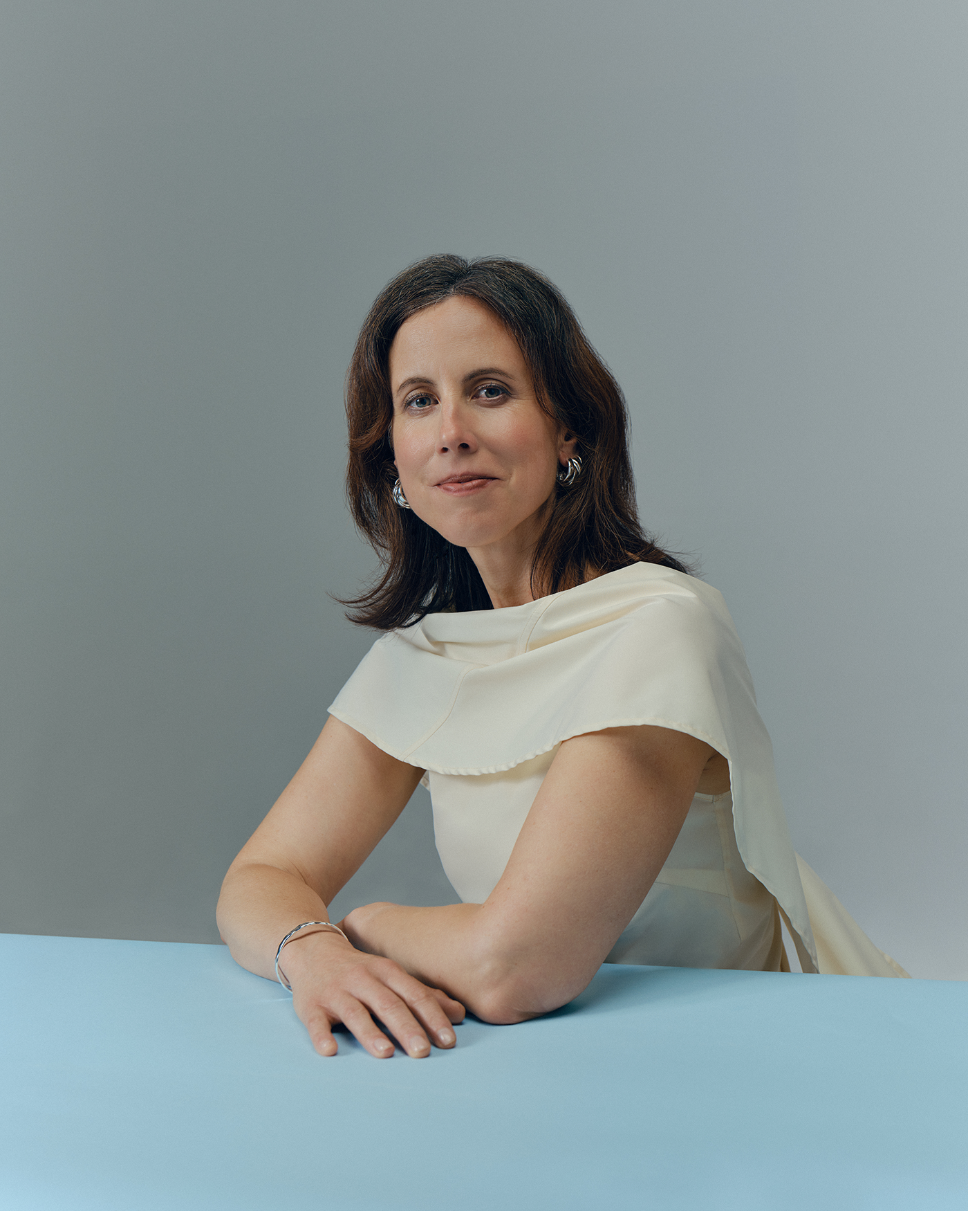 A woman in an ivory outfit leans forward with her arms resting on a light blue table against a plain grey background.