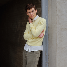 Sam Nivola photographed by JJ Geiger. He is leaning against a cement wall with his arms crossed wearing a butter yellow cardigan over a white down down shirt and gray jeans. 