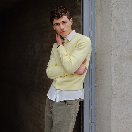 Sam Nivola photographed by JJ Geiger. He is leaning against a cement wall with his arms crossed wearing a butter yellow cardigan over a white down down shirt and gray jeans. 