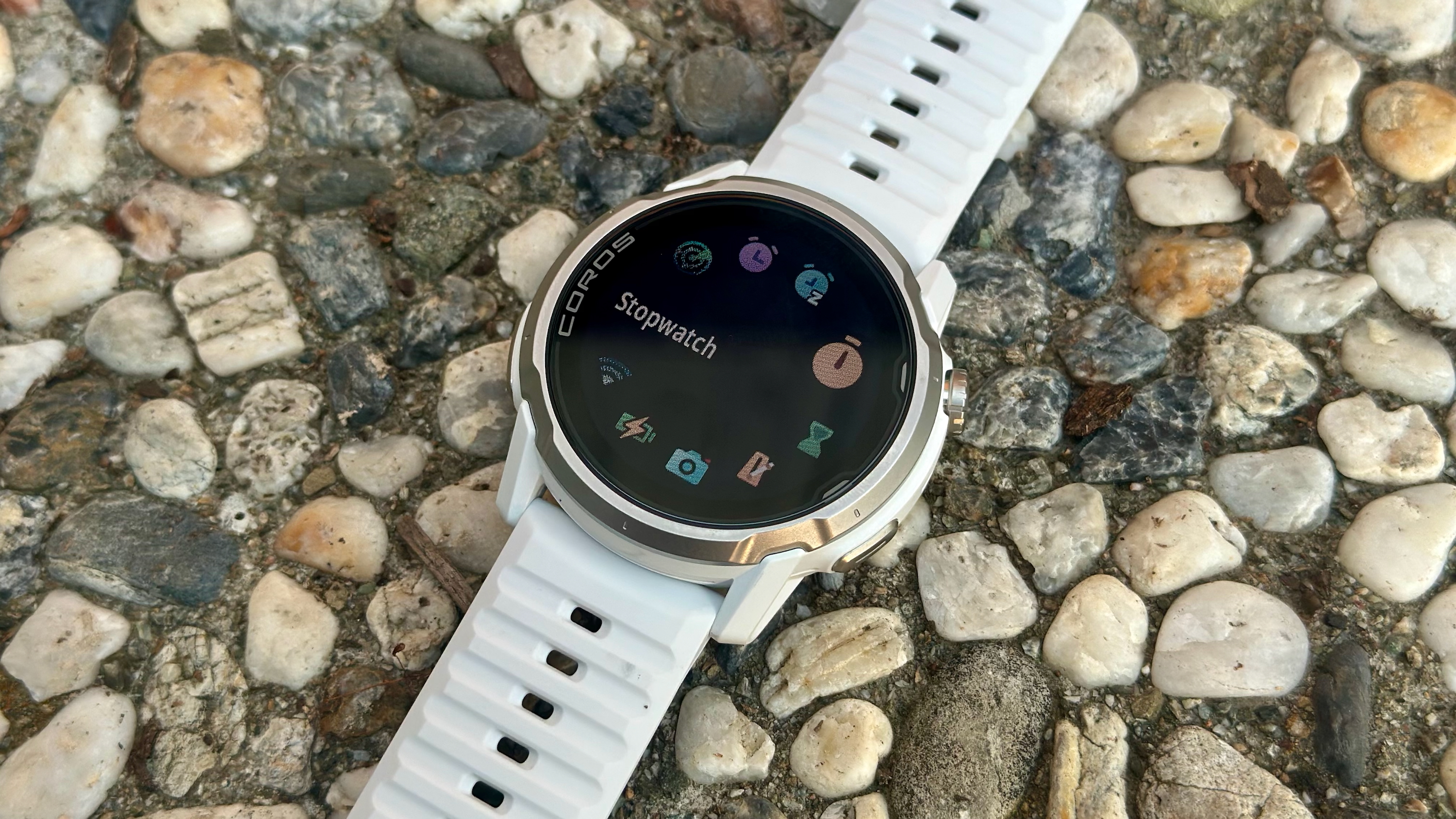 A photo of the COROS APEX 4 sitting on a rocky surface, showing the Toolbox menu with the Stopwatch app highlighted and other icons visible in a circle.