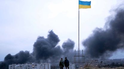 People in front of an Ukrainian national flag watching dark smoke rise following an air strike
