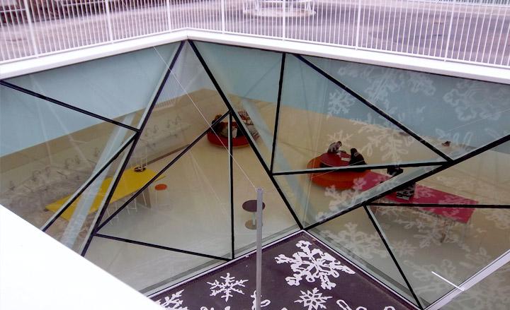 View into underground space looking through glass ceiling constructed of triangular panes of glass with black trims.