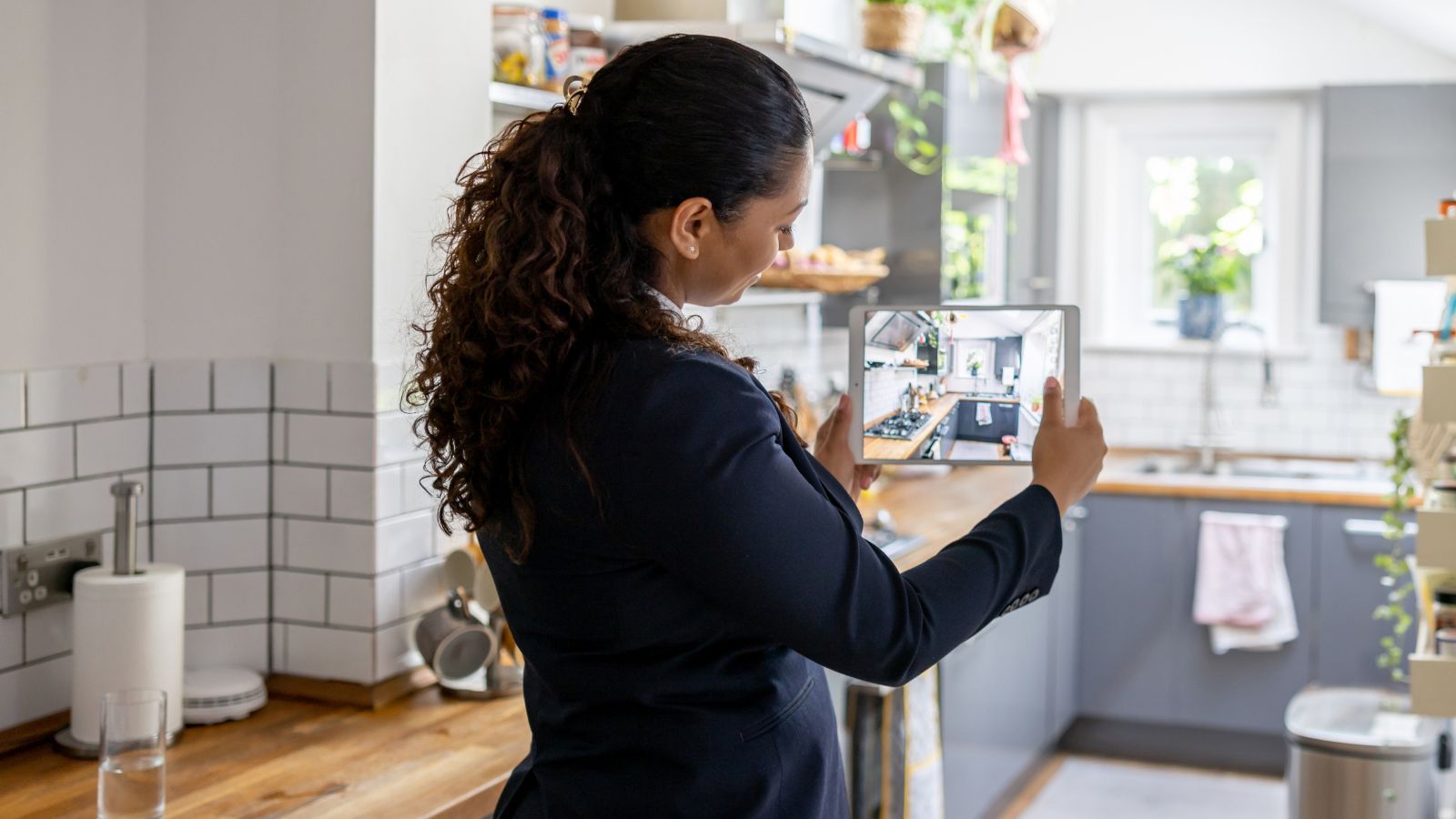 A dark-haired woman in a navy blazer photographs a grey and wood kitchen on a tablet, likely an estate agent conducting a property valuation.
