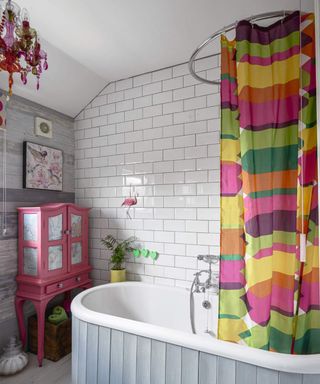 Bathroom with shower over bathtub with multicolored shower curtain and chandelier with white subway wall tiles and pink armoire