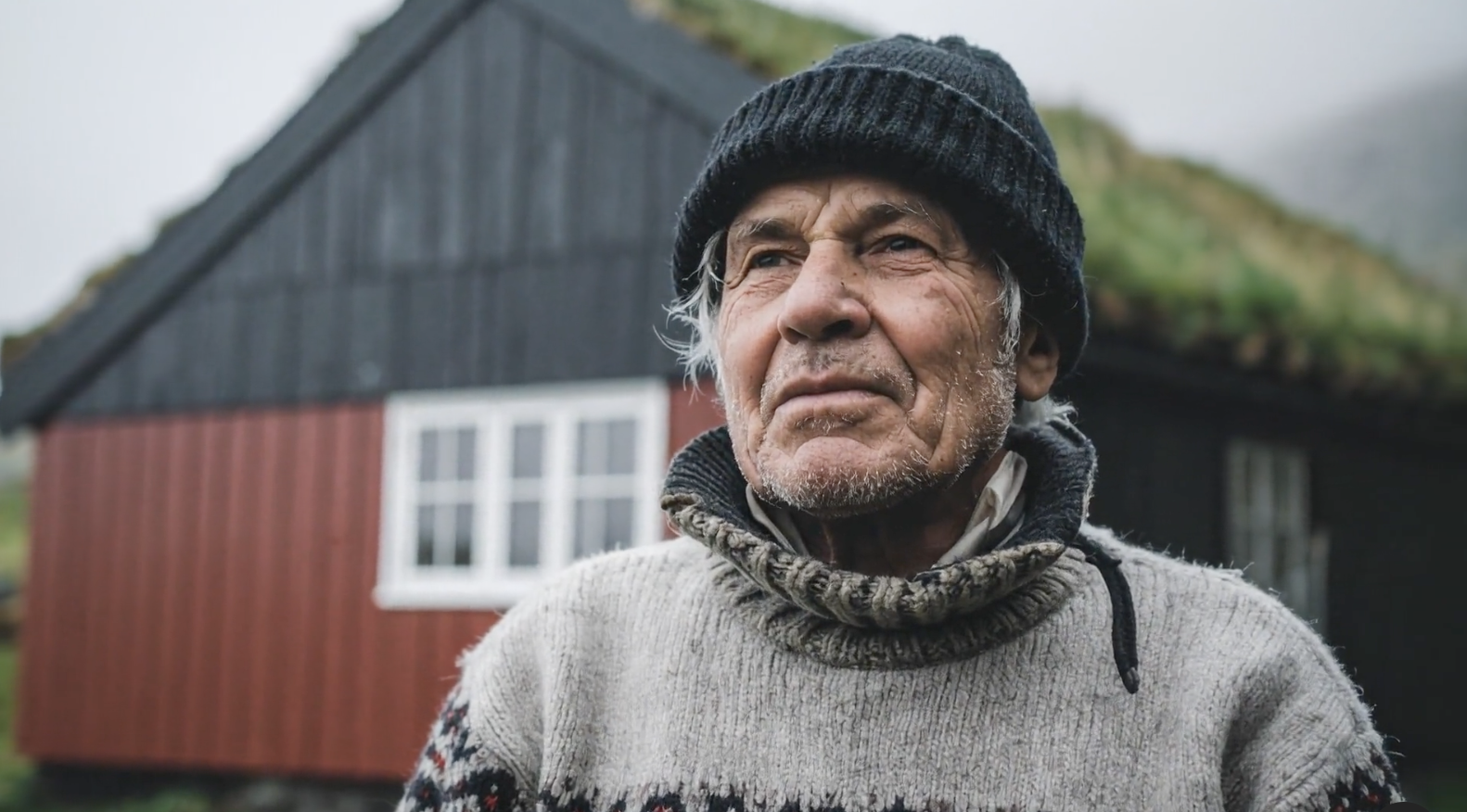 A man with a weathered face and a knit cap looks thoughtfully to the right, standing outdoors in front of a rustic building with a dark roof and red siding.