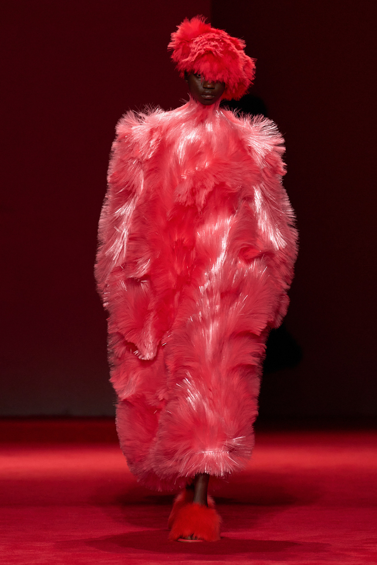 A model walking in the Bottega Veneta F/W 26 show wearing a hot pink fringe coat with a red fringe hat and red furry shoes.