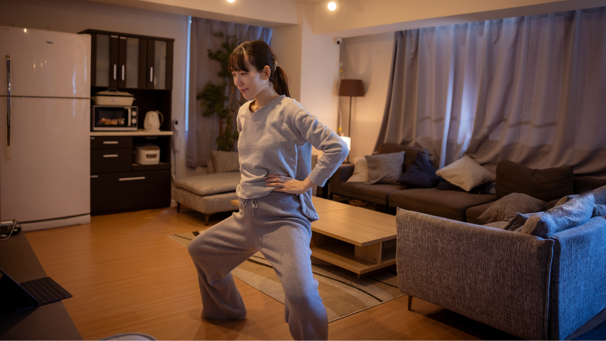 Woman standing in a domestic living area in an athletic stance with her hands on her hips, feet wider than shoulder-width apart and knees bent