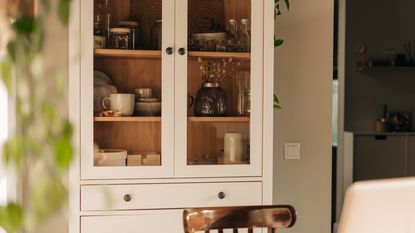 An image of a white kitchen cabinet with glass doors and a selection of crockery inside, with a wooden table in the foreground and soft focus foliage on left