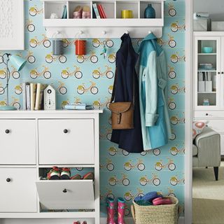 A white shoe storage unit in a hallways with turquoise patterned wallpaper and a wall shelf