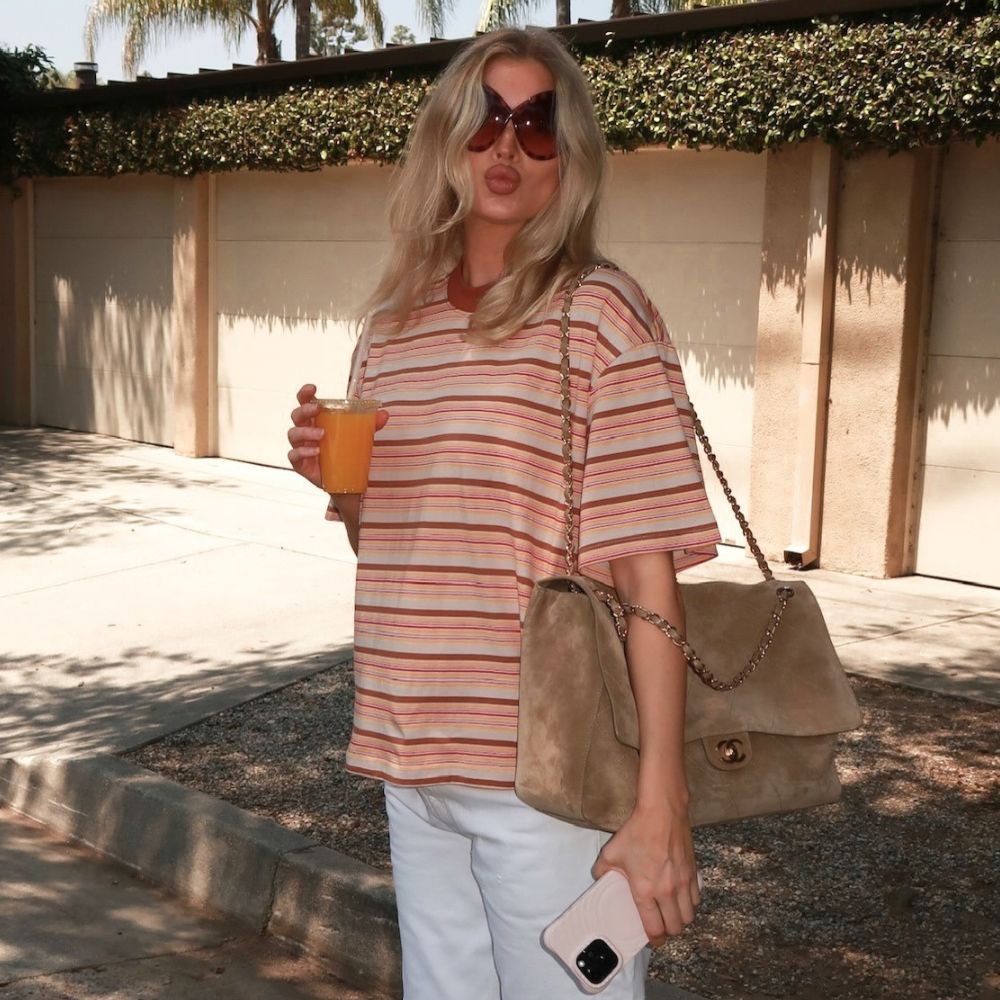 Elsa Hosk holds an orange juice wearing a striped t-shirt with white jeans and brown flip-flops.