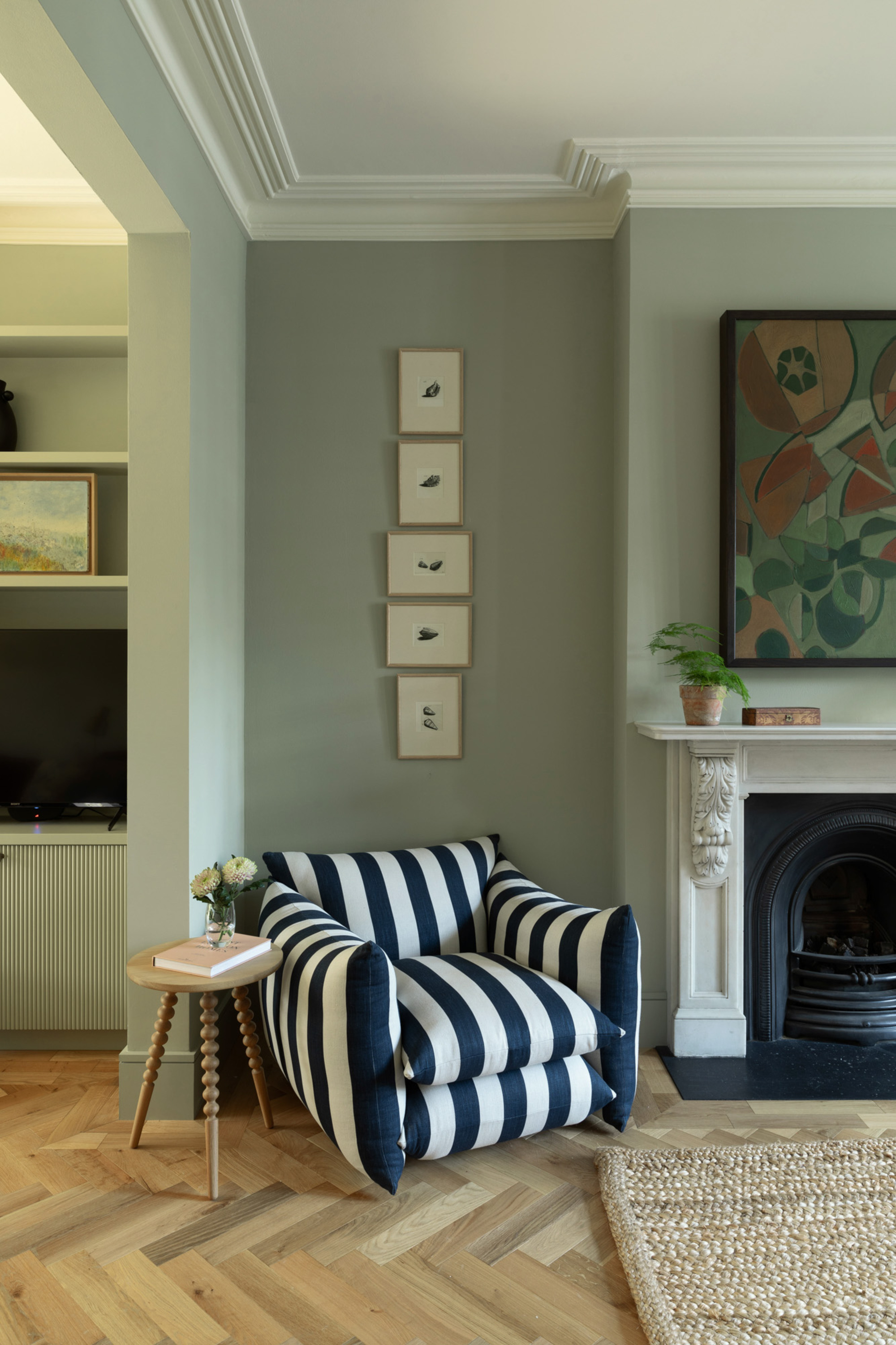 living room with sage green walls, timber parquetry floors, a black and white striped armchair with a bobbin side table, framed artworks, an ornate fireplace with a jute rug in front