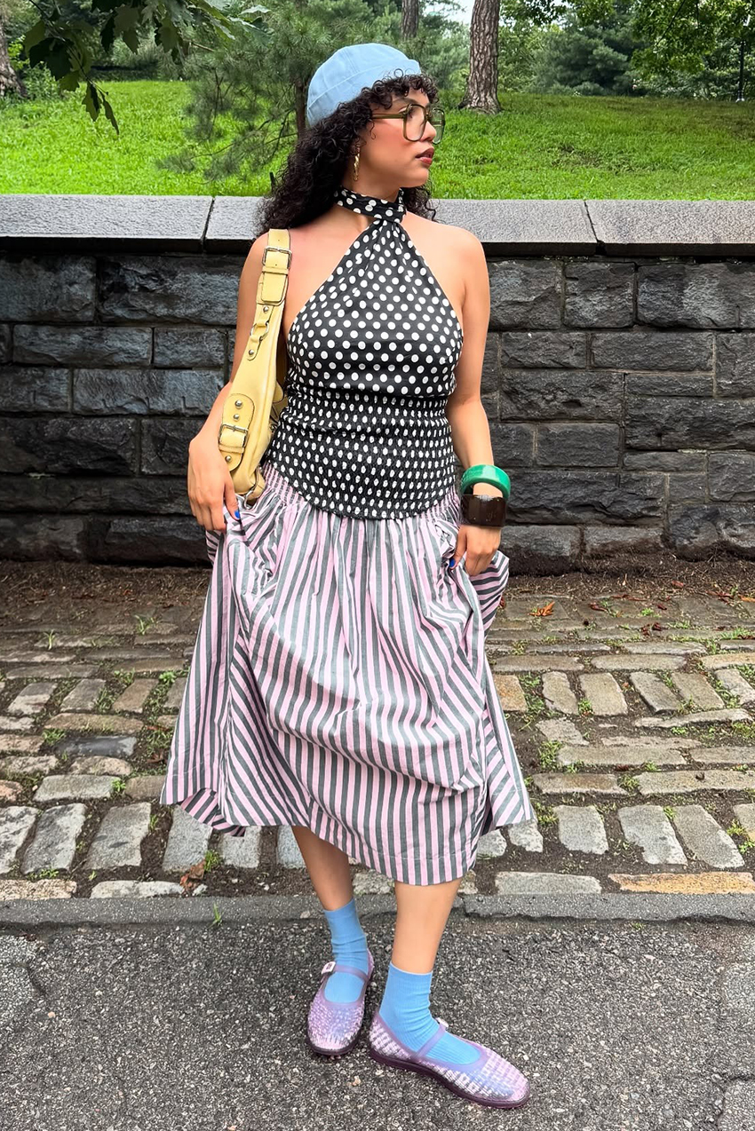 a woman wearing a blue hat, polka dot shirt, striped skirt, blue socks, and lavender flats