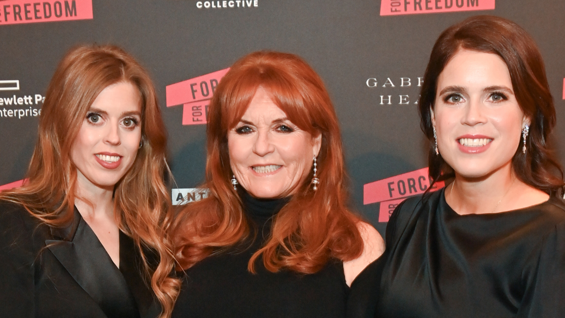 Princess Beatrice, Sarah Ferguson and Princess Eugenie posing in black dresses on the red carpet in front of a black step and repeat reading "Freedom"