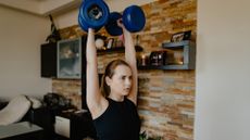 A woman doing a dumbbell standing workout in her living room, performing the overhead press