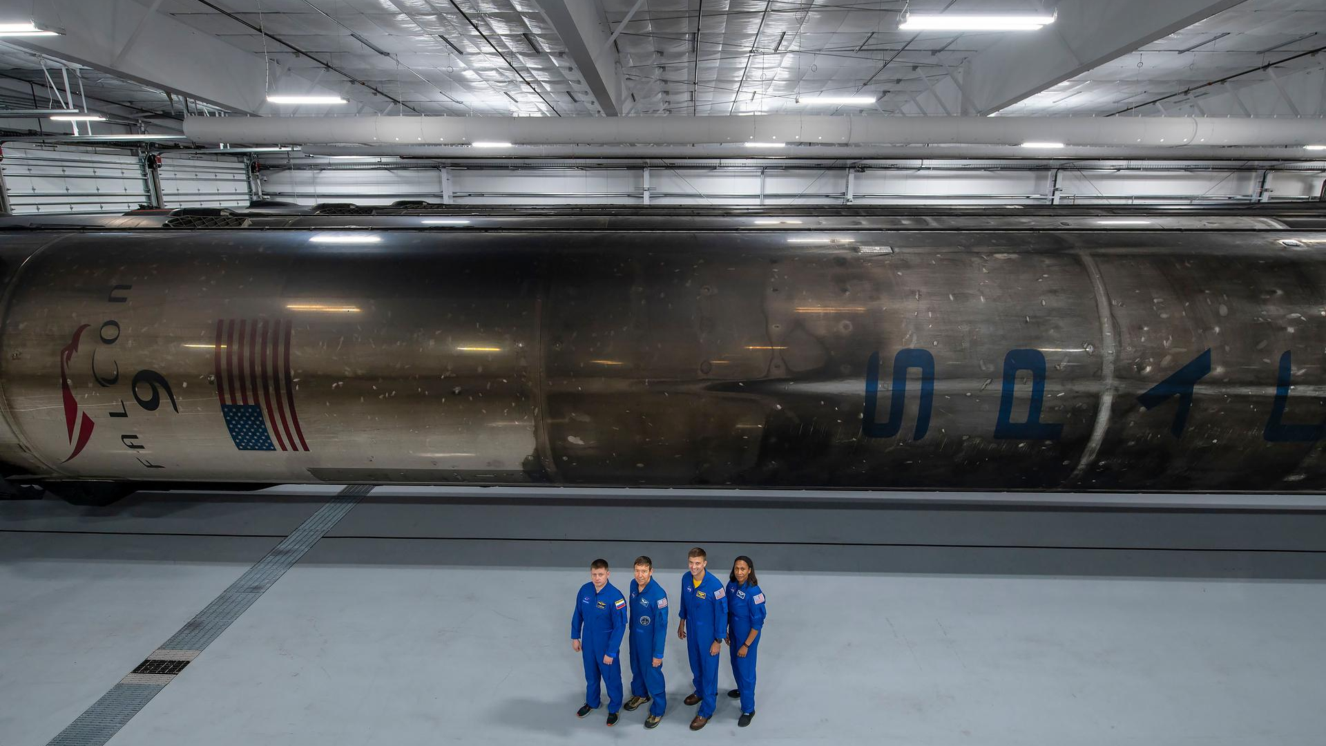 four humans in blue jumpsuits stand in a large white hanger with a relatively low ceiling. Behind them, laid on its side, is a sooty rocket booster, smeared in black from the char returning through earth's atmosphere during previous flights.