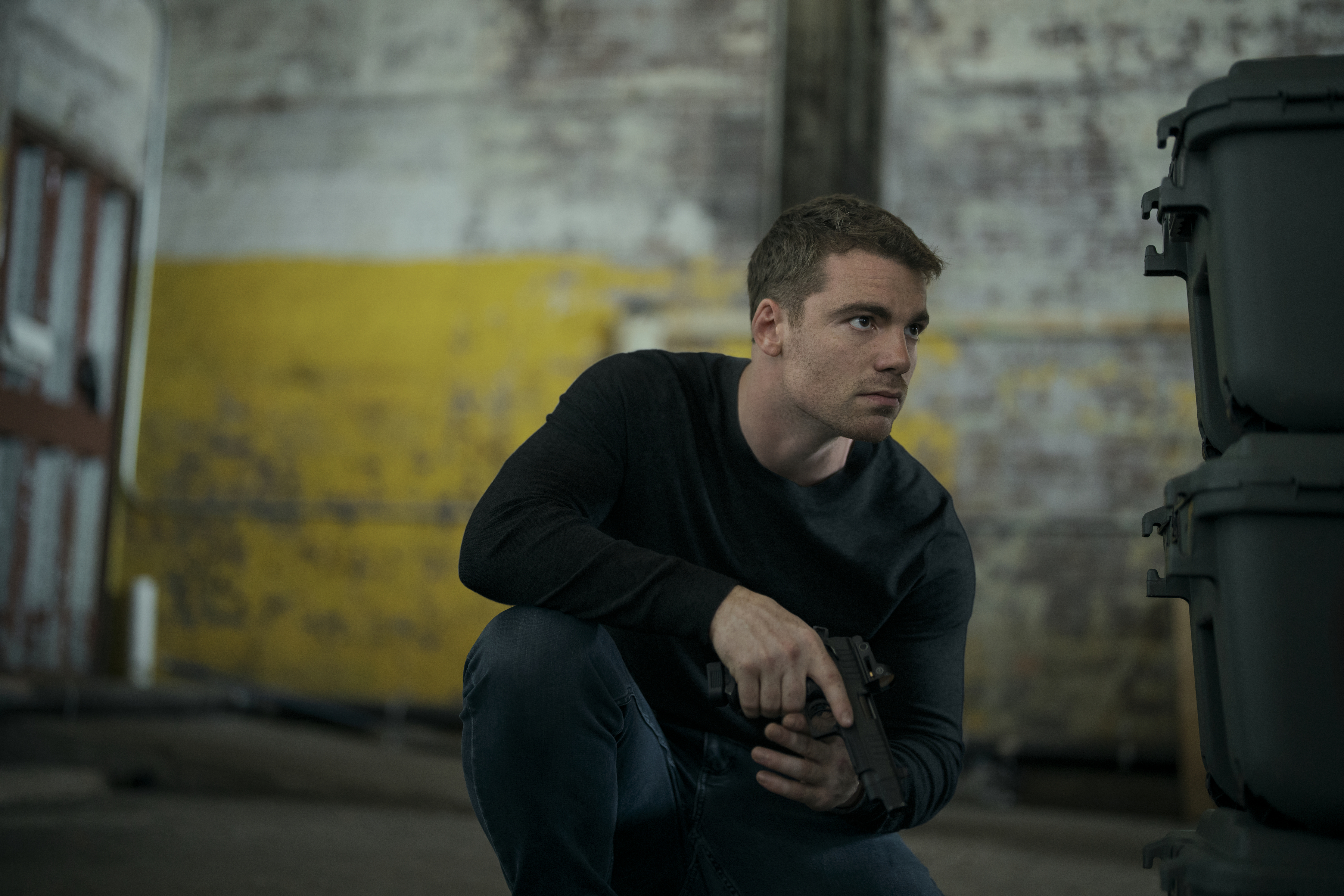 Gabriel Basso as Peter Sutherland in episode 309 of The Night Agent hiding behind bins in a warehouse getting ready to shoot a gun