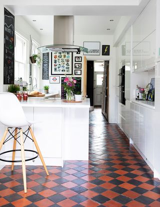 A white kitchen with island, bar stool, framed wall art and terracotta and black checkered flooring