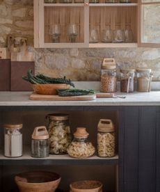 A rustic kitchen with white countertops. Glass jars of dried food goods on the counter and shelves beneath. Dark blue pained cabinets, 