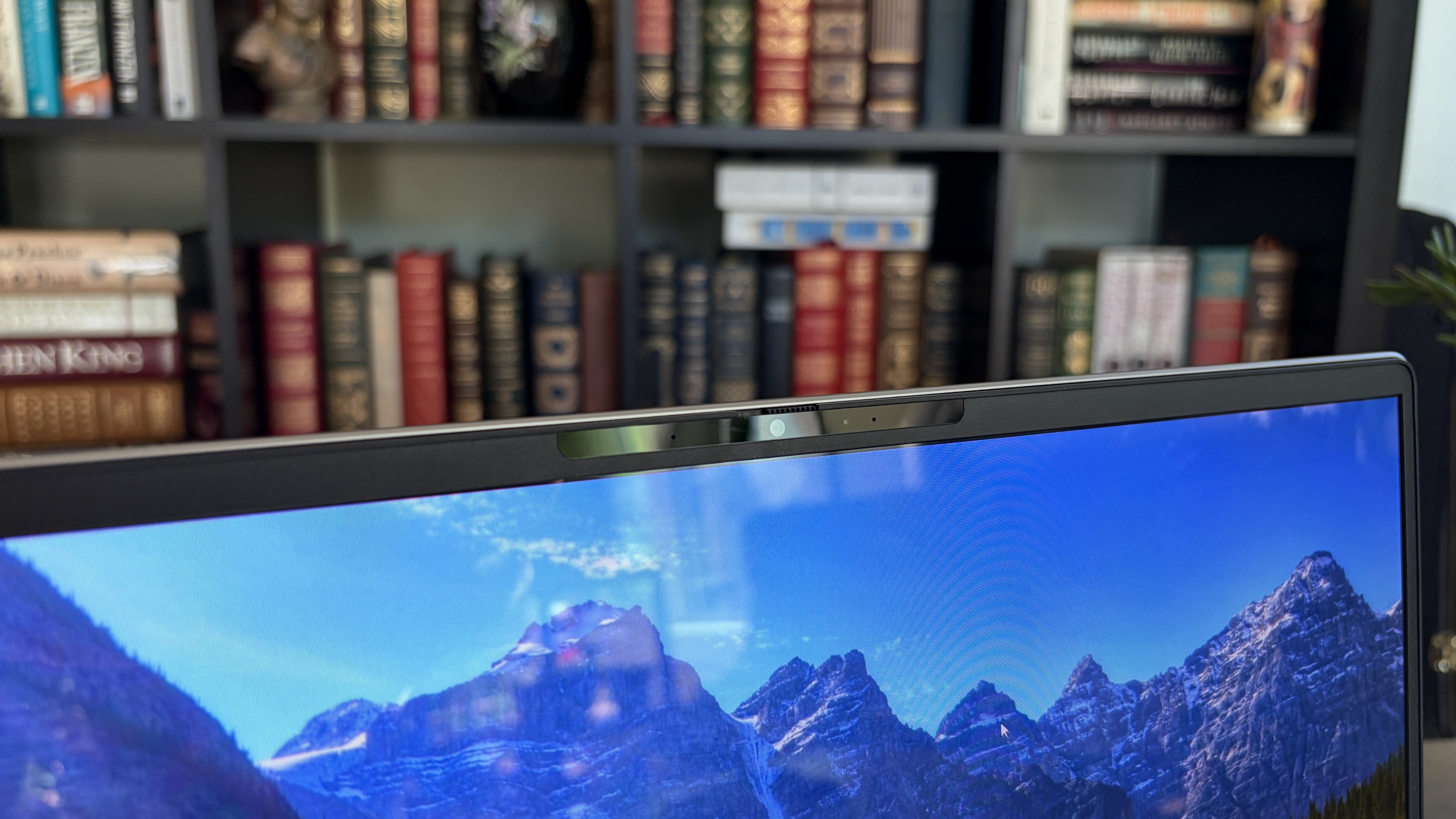 Close-up of a laptop screen with a built-in webcam, displaying a scenic mountain landscape, and a bookshelf filled with colorful books in the background.