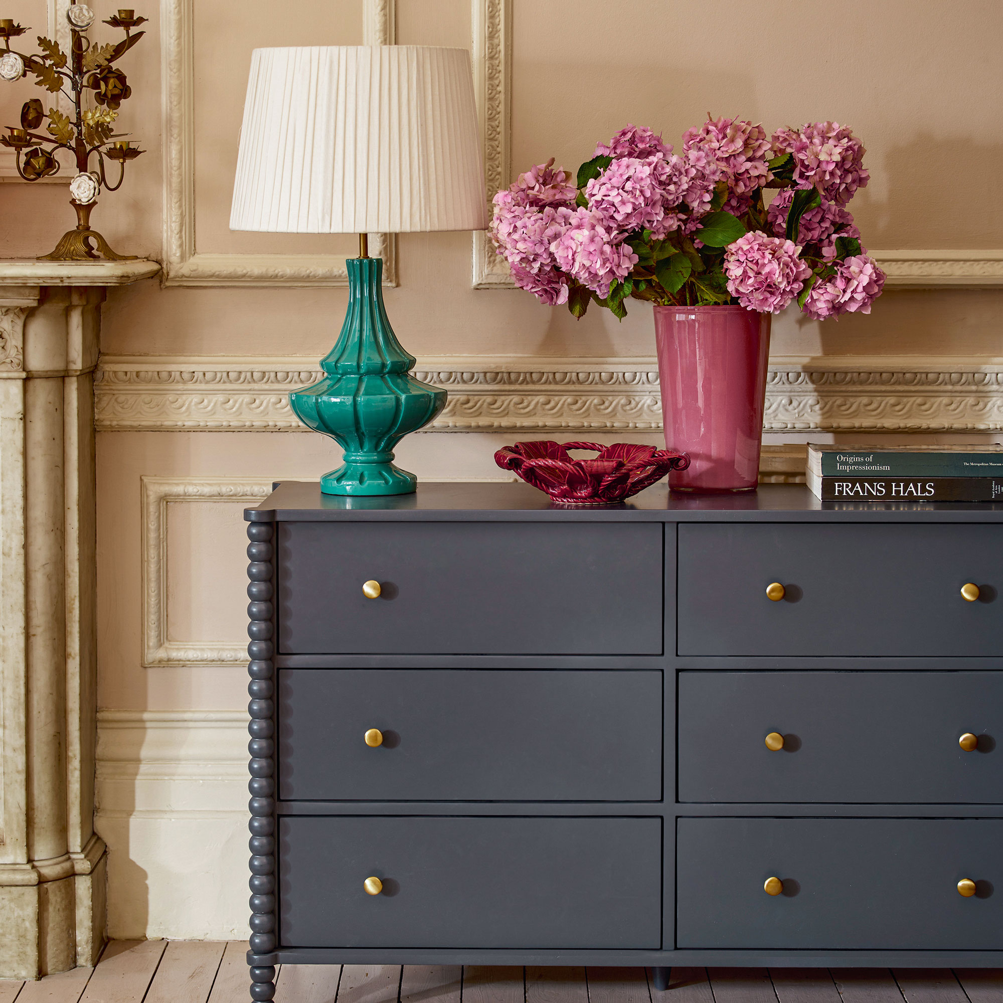 White and pink ornate living room with a navy chest of drawer, blue lamp and pink vase filled with hydrangeas