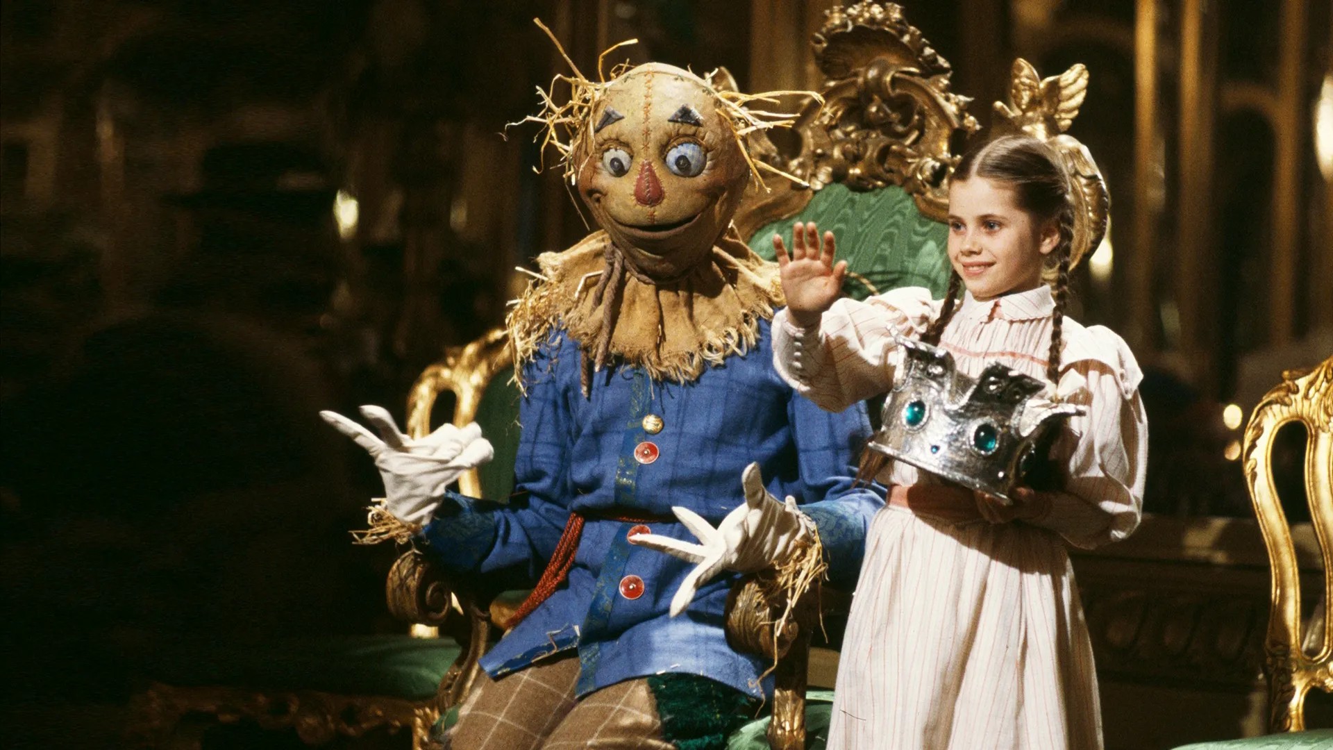 A still from Return to Oz showing Dorothy, in a striped dress and plaited hair, holding a jewelled silver crown beside the Scarecrow on an ornate gilded throne.
