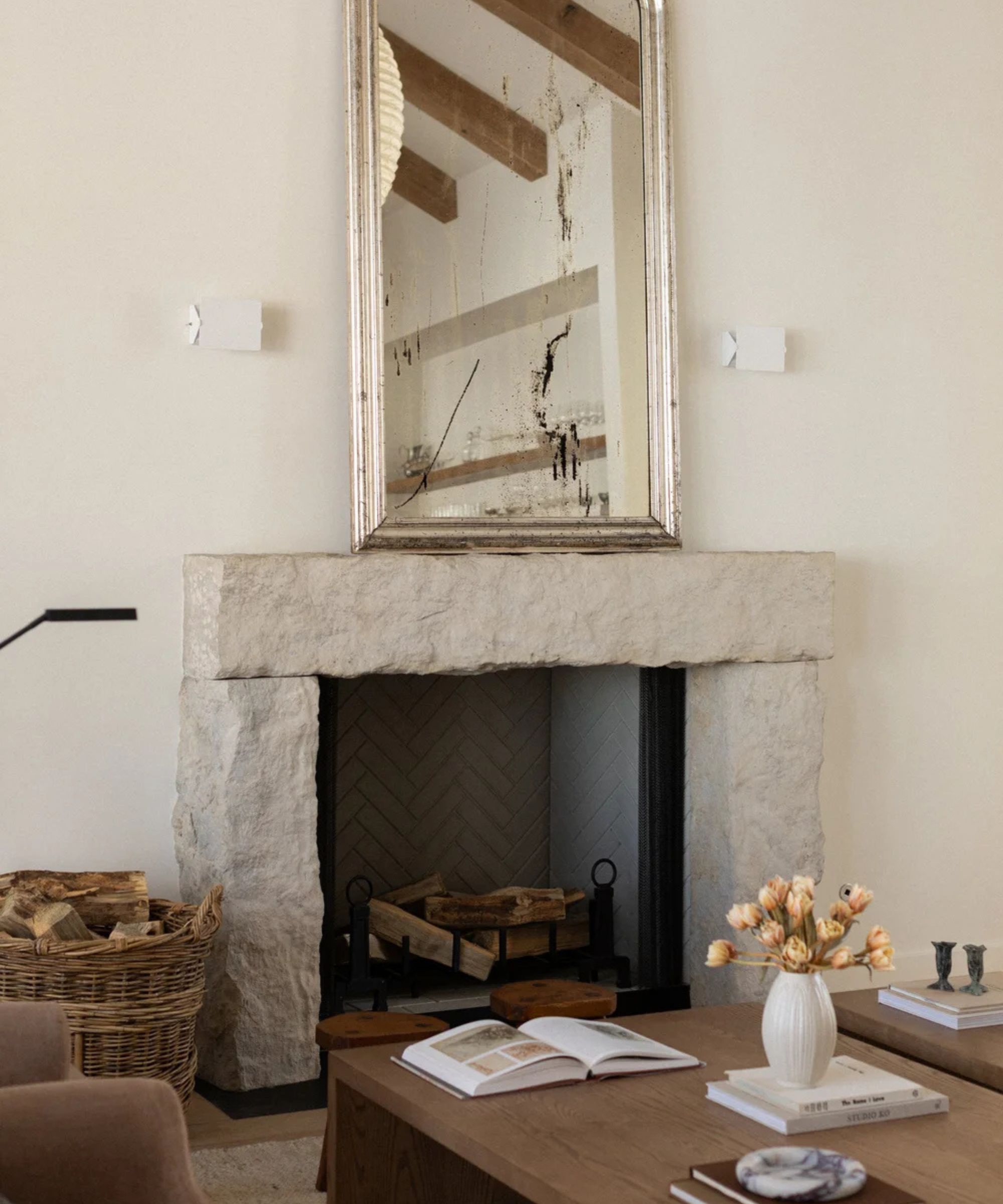 Reclaimed stone fireplace surround with a chunky, textured mantel beneath a distressed antique mirror, styled with a woven log basket, stacked firewood and a wooden coffee table topped with books and a vase of tulips.