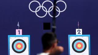 The back of the head of an archer is shown as they aim at the target. Two targets are shown, one on the left is numbered with a 1 and another on the right is numbered with a 2. Between the two targets are the olympic rings in white against a navy blue background.