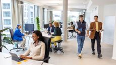 Workers in an open plan office space using AI on desktop computers while others work on laptops, with colleagues walking to a meeting.