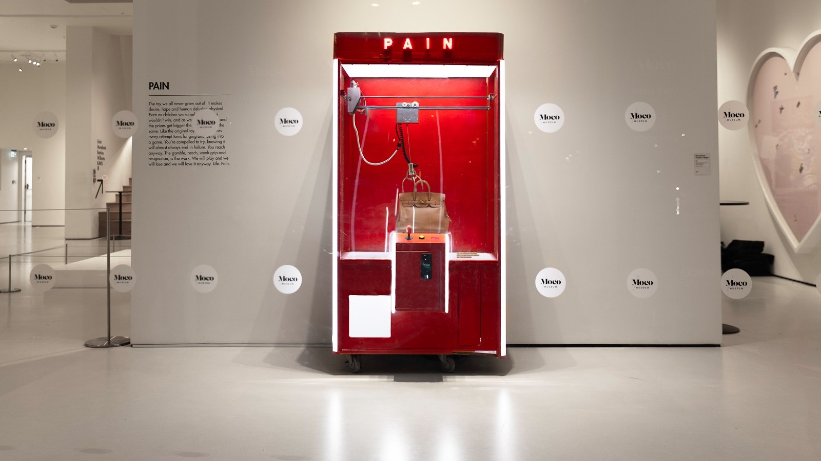 A brightly lit art installation at the Moco Museum features a red claw crane machine labeled "PAIN" containing a single tan luxury handbag.