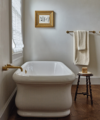 white bathtub in simple bathroom with wooden floors and stool with soap and towel rail and art in background on wall