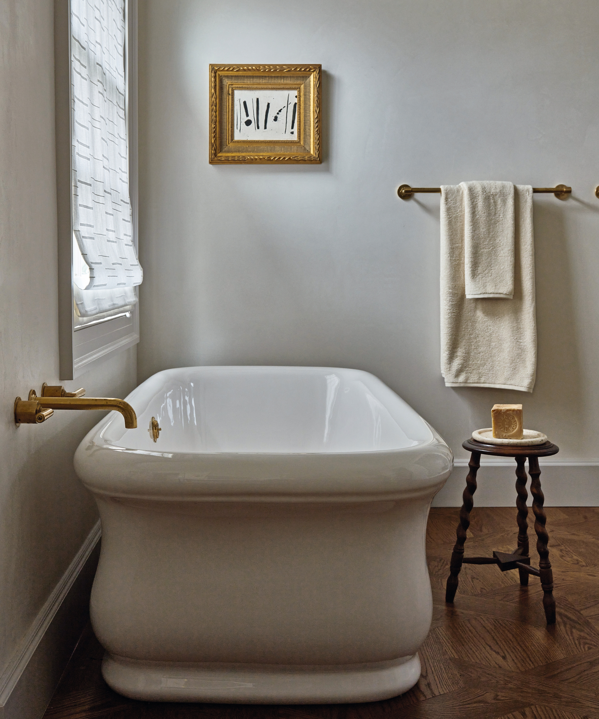 Baignoire blanche dans une salle de bains simple avec parquet et tabouret avec porte-savon et porte-serviettes et œuvres d'art en arrière-plan sur le mur